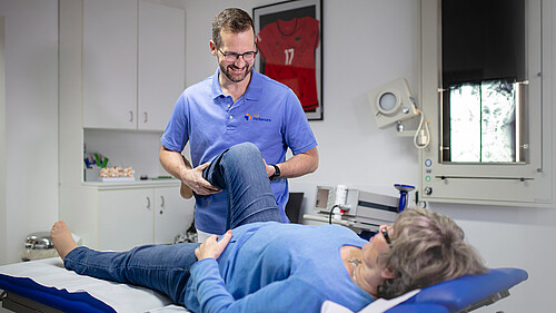 Arzt untersucht das angewinkelte Knie einer Patientin auf der Behandlungsliege im Behandlungszimmer.