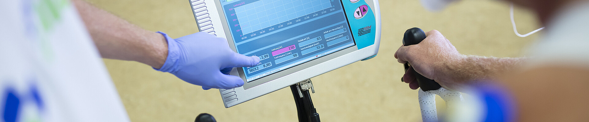 Perspektive über die Schulter einer Person, die eine Leistungsdiagnostik durchführt. Im Bild ist ein Sportwissenschaftler zu sehen, der den Sportler betreut, sowie ein Bildschirm, auf dem die Werte der Diagnostik angezeigt werden.